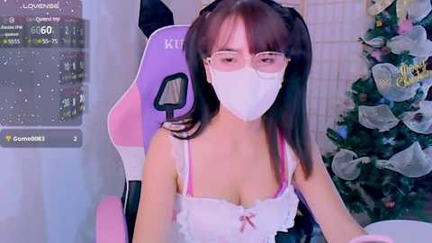 Media: A video of a young Asian woman with glasses and a face mask, wearing a white lacy top, sitting in a pink gaming chair, surrounded by a festive Christmas tree.