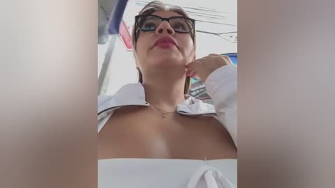 Media: A video shows a woman with glasses and a white blouse, partially open, revealing cleavage, standing in a public place. The background includes blurred objects and electrical wires.