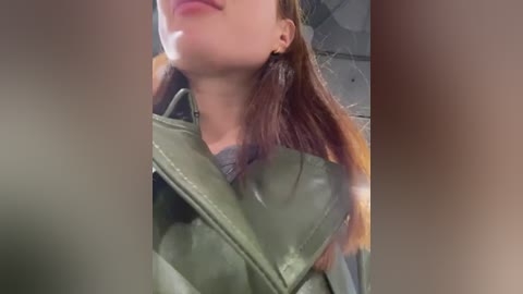 Media: Video of a young woman with light skin, wearing a green leather jacket, brown hair, and minimal makeup, partially obscured by a blurred background.