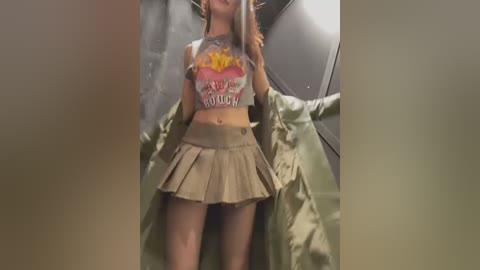 Media: Video of a young woman with light skin and long brown hair, wearing a cropped, graphic T-shirt and a short, pleated tan skirt, standing in a dimly lit, cluttered room with metal objects and a green jacket draped over her shoulders.