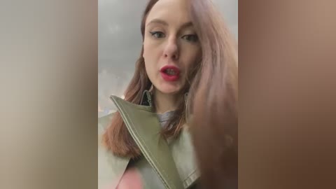 Media: A video of a young woman with fair skin, straight brown hair, and red lipstick, wearing a green jacket, looking shocked and pointing at something off-screen.