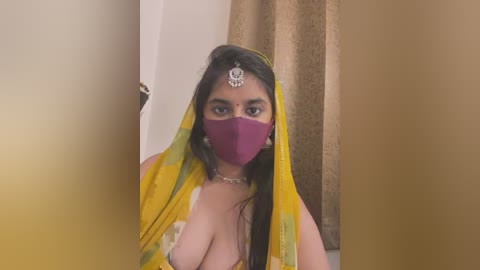 Media: Video of a dark-skinned woman with long black hair wearing a yellow saree, purple face mask, and a forehead bindi, set against a beige curtain background.