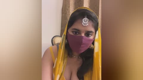 Media: Video of a young South Asian woman with medium skin tone, wearing a yellow saree, purple mask, and ornate headpiece, against a beige and brown backdrop.