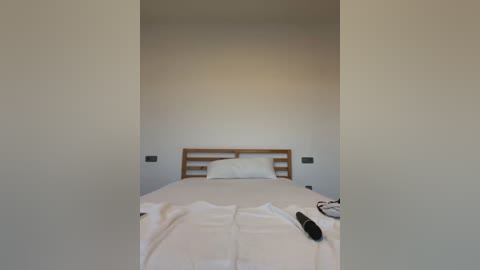 Media: Video of a minimalist bedroom with a wooden headboard, white bed linens, and a remote control on the bed. The room has plain white walls and two small black outlets on either side of the headboard.