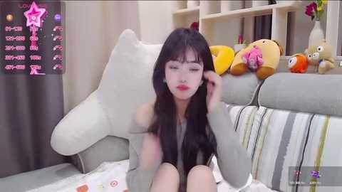 Media: Video of an Asian woman with long black hair and fair skin, sitting on a gray couch, surrounded by plush toys and a pink star logo in the corner.