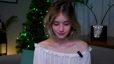 Media: Video of a young blonde girl with shoulder-length hair, wearing an off-the-shoulder white dress, smiling, standing in a cozy living room with a lit Christmas tree and potted plants.