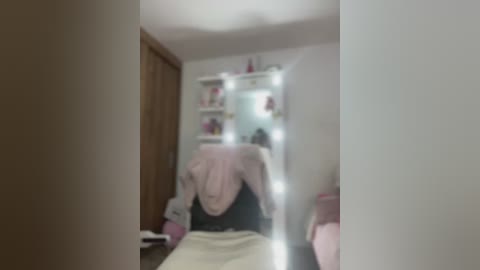 Media: Blurry video of a bedroom with a white bed, pink blanket, and a white vanity mirror with light bulbs, reflecting a girl wearing a black shirt.