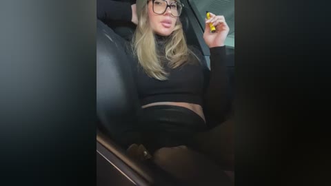 Media: Video of a blonde woman with glasses, wearing a black crop top and high-waisted pants, sitting in a car with a yellow object in her hand, looking confident.