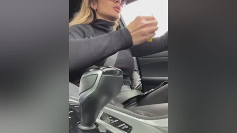 Media: Video of a blonde woman in a black turtleneck, driving a car with a visible black steering wheel, holding a yellow pen, and wearing sunglasses.