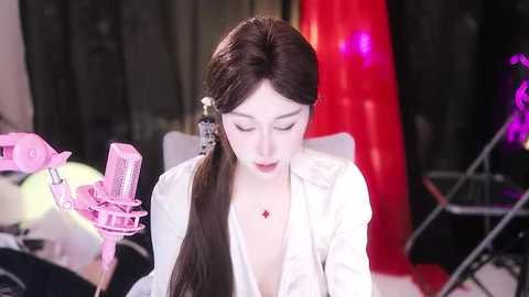 Media: A video of a young Asian woman with long dark hair, wearing a white blouse and red star necklace, using a pink microphone to record herself. The background features a red curtain and studio equipment.