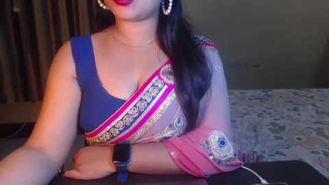 Media: Video of a woman with medium skin tone, wearing a blue blouse and pink saree with intricate embroidery, seated at a table. She has long dark hair and wears large pearl earrings.