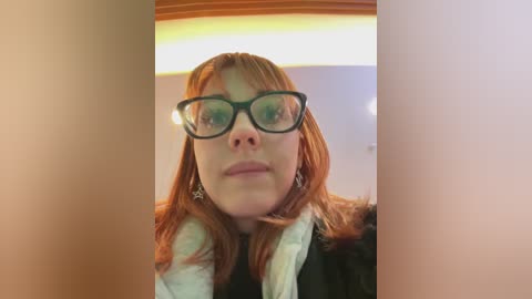 Media: Video of a fair-skinned woman with long red hair, wearing black-framed glasses, a white scarf, and a black jacket, indoors with warm lighting.