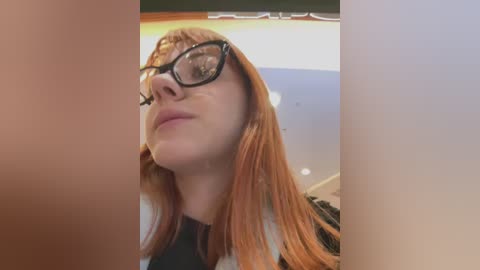 Media: Video of a woman with long, straight, auburn hair, wearing black-rimmed glasses, looking upwards with a neutral expression, in a dimly lit room with white walls and a wooden ceiling.