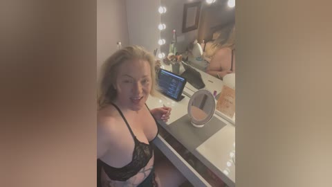 Media: Video of a smiling, fair-skinned woman with shoulder-length blonde hair, wearing black lace lingerie, sitting at a makeup vanity with a mirror, surrounded by personal items and a laptop.