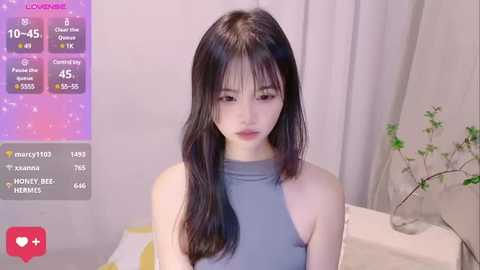 Media: Video of a young East Asian woman with long black hair, wearing a sleeveless light blue top, sitting indoors with a minimalistic background.