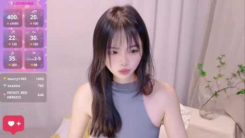 Media: Video of an East Asian woman with long, dark hair, wearing a sleeveless grey top, sitting on a bed with a potted plant. The background features a beige curtain and a digital health tracker screen.