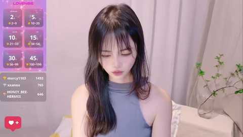 Media: Video of an East Asian woman with long, straight black hair, wearing a sleeveless gray top, indoors. Background includes a bed with white sheets and a potted plant.