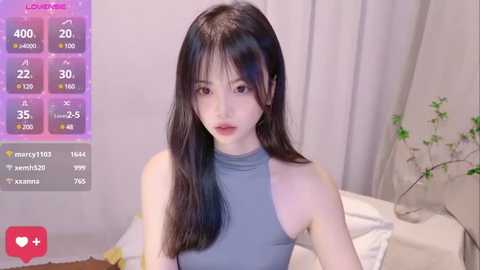 Media: Video of an East Asian woman with long black hair, wearing a sleeveless, high-necked gray top, sitting on a bed with white sheets and a potted plant in the background.