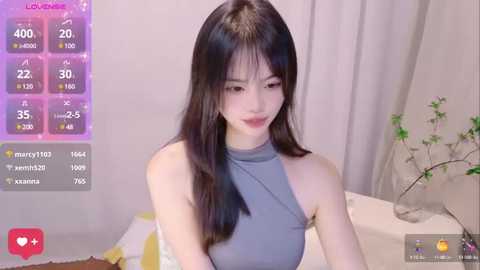 Media: Video of a young East Asian woman with long black hair, fair skin, and a slim figure, wearing a grey halter top. Background features a white curtain and a vase with green plants. Social media metrics are overlaid.