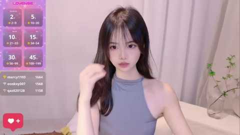 Media: Video of an Asian woman with long, straight black hair and fair skin, wearing a sleeveless gray top, sitting on a white couch. Background features a soft purple and white gradient, a plant, and a \"Love Live!\" chart.