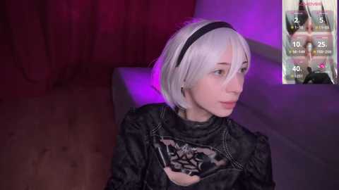 Media: Video of a pale-skinned woman with short, platinum-blonde hair, wearing a black and white cosplay of 2B from \"Nier: Automata,\" sitting in a dimly lit room with red and purple lighting.