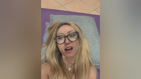 Media: Video of a blonde woman with glasses and a necklace, lying on a purple yoga mat with a gray pillow behind her head, in a room with a tiled floor.