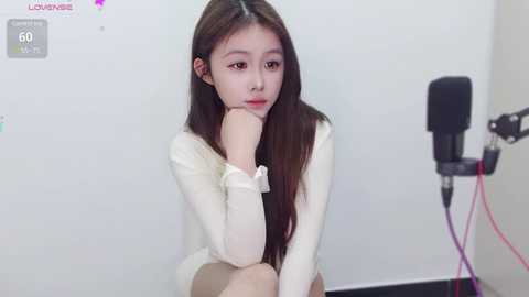 Media: Video of an East Asian woman with long brown hair, wearing a white sweater, sitting pensively against a white wall, with a microphone stand and camera on the right.