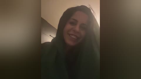 Media: Video of a smiling, dark-haired woman with fair skin, partially obscured by a dark curtain. Soft lighting creates a warm, intimate atmosphere.