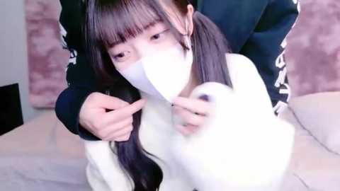 Media: Video of a young Asian woman with long black hair in pigtails, wearing a white face mask, holding a fluffy white stuffed animal, in a room with a pink plush pillow and light-colored bedding.