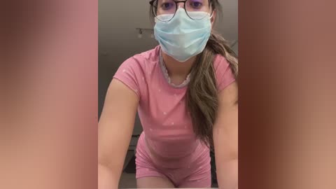 Video of a young woman in a pink T-shirt and matching shorts, wearing a blue surgical mask and glasses, leaning forward, with blurred people's legs in the background.