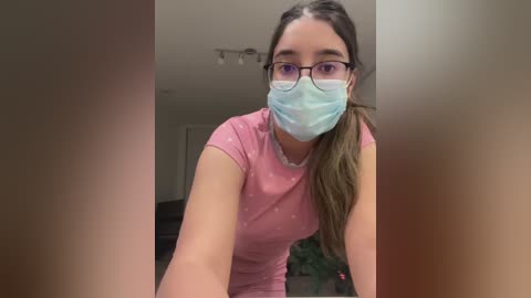 Video of a young woman with glasses, light skin, and a light pink t-shirt, wearing a blue surgical mask, taken indoors.