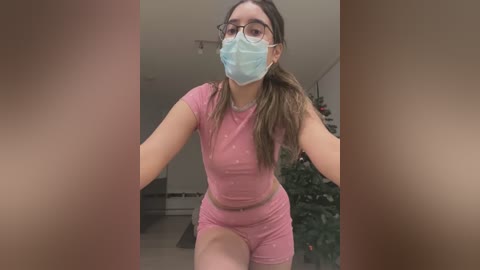 Media: Video of a young woman with light skin, brown hair, and glasses, wearing a pink crop top and shorts, a face mask, and a festive background with a decorated Christmas tree.