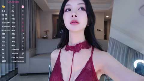 Media: Video of an Asian woman with long black hair, fair skin, and full lips, wearing a red lace bralette, standing in a modern, dimly lit living room with grey curtains and a couch.