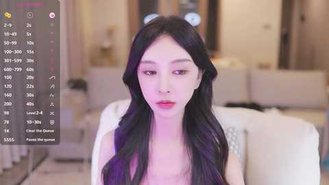 Media: Video of a fair-skinned East Asian woman with long, wavy black hair, wearing a white top, in a modern, softly lit living room with neutral tones and a plush white couch.