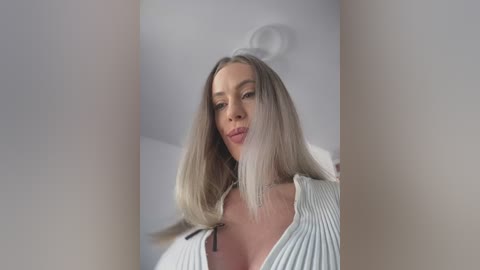 Video of a light-skinned woman with long, straight blonde hair and a deep V-neck white sweater. She has a soft, serene expression, set against a minimalist, softly lit background.