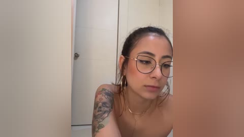 Media: Video of a young Latina woman with light brown skin, wearing glasses and a gold necklace, looking into a mirror. She has a tattoo on her left arm, and the background shows a beige bathroom door.