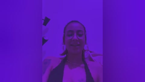 Media: A video of a smiling woman with light skin and brown hair, wearing a black bikini top, standing in a room bathed in purple light.