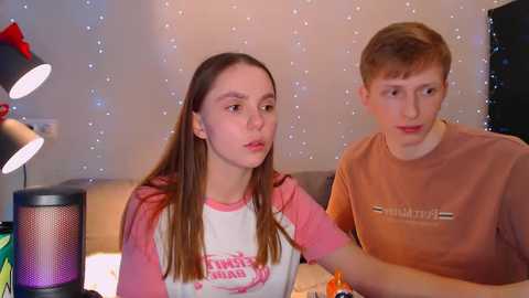 Media: Video of a young white girl with long brown hair in a pink and white baseball shirt, and a boy with short red hair in a brown shirt, seated at a table, indoor setting with string lights, lamps, and a speaker.