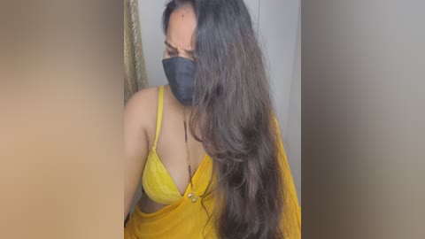 Media: A video of a South Asian woman with long, dark hair, wearing a yellow saree, black mask, and a black bra, leaning against a beige wall.