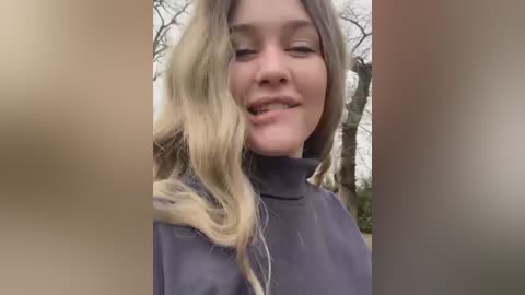 Media: Video of a young Caucasian woman with wavy, blonde hair, wearing a dark purple turtleneck sweater, smiling outdoors with blurred background of leafless trees.