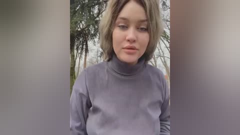 Media: Video of a young woman with shoulder-length, wavy blonde hair, wearing a gray turtleneck sweater, standing outdoors with a blurred background of trees and foliage.