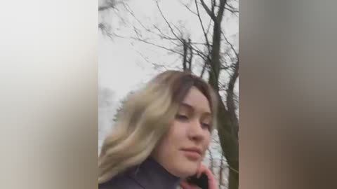 Media: Video of a fair-skinned young woman with long, wavy blonde hair, wearing a dark coat, standing in a wintry forest with bare trees.