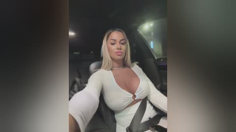 Media: Video of a blonde woman with medium skin tone, wearing a low-cut, white crop top, driving a car at night.