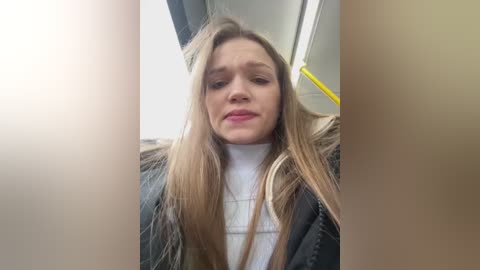 Media: Video of a blonde woman with straight hair, wearing a black leather jacket over a white turtleneck, sitting inside a public bus, with a yellow pole visible in the background.