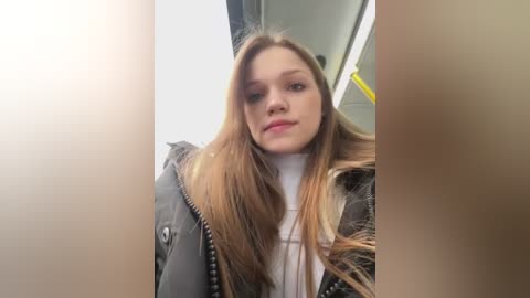 Media: A video of a young woman with long, straight, light brown hair and pale skin, wearing a dark coat and white turtleneck sweater, taken inside a bus with yellow handles visible.