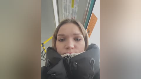 Media: Video of a young woman with straight, light brown hair, wearing a black puffy jacket, partially covering her face. She's standing in a subway station with yellow handrails and a blurred background.