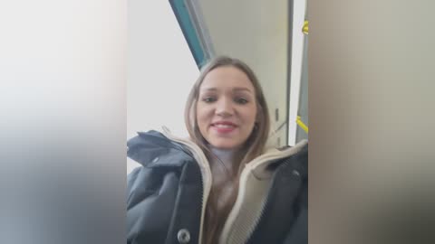 Media: A video of a young, fair-skinned woman with long, straight brown hair, wearing a black and beige puffer jacket, smiling. The background shows a subway car interior with blue seats.