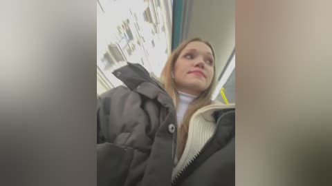 Media: A video of a young woman with fair skin and long, straight blonde hair, wearing a grey coat and white turtleneck sweater, looking contemplative while riding a subway train.