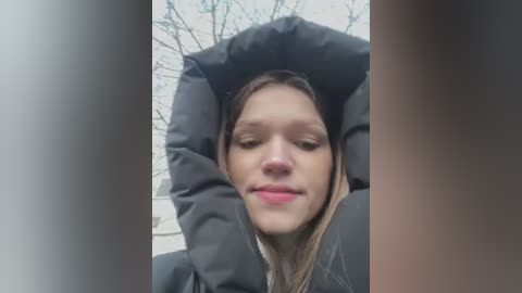 Media: Video of a young Caucasian woman with light skin and blonde hair, wearing a black puffy jacket, sitting outdoors with bare tree branches in the background.