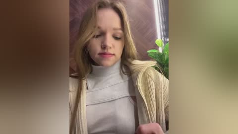 Media: Video of a young Caucasian woman with light brown hair, wearing a cream-colored turtleneck and beige cardigan, sitting with eyes closed and a contemplative expression, in an indoor setting with a wooden background and a potted plant visible.